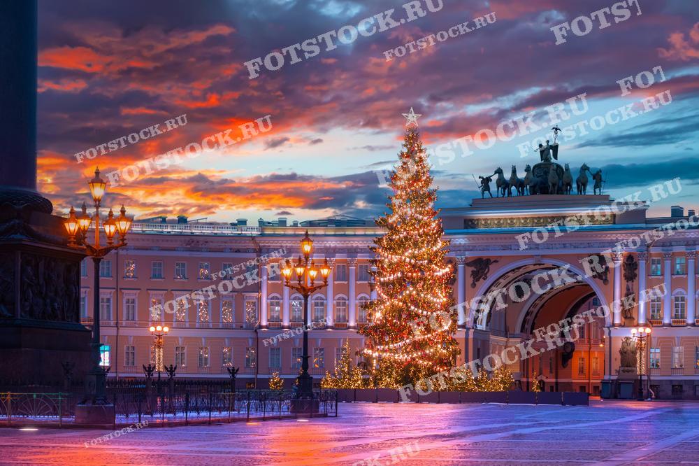 На фотографии изображена Дворцовая площадь в Санкт-Петербурге зимой, украшенная праздничной елкой. Вечернее небо окрашено в теплые оттенки заката, создавая живописный фон. Здание Главного штаба с его величественной аркой и иллюминацией добавляет торжественности. Этот вид передает атмосферу праздника и волшебства зимнего Петербурга.